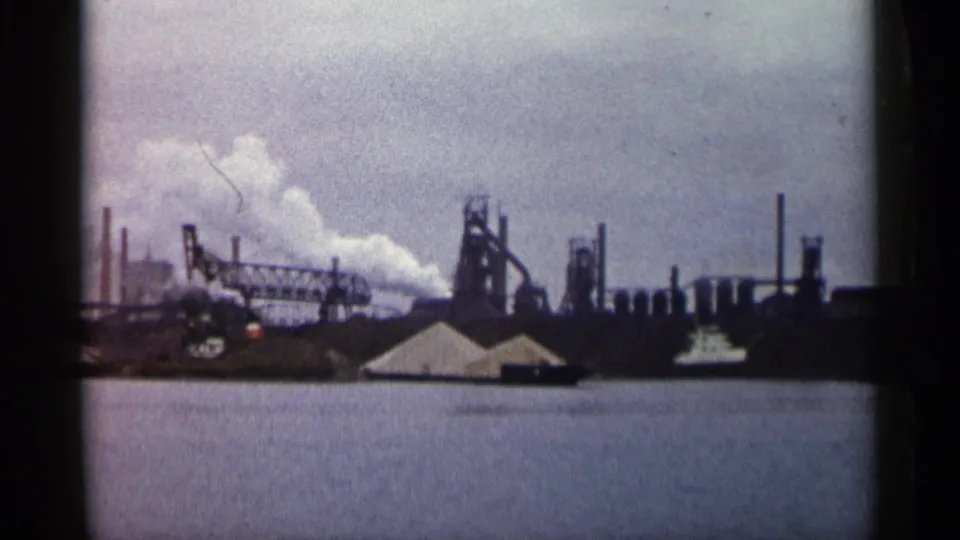 1960 Michigan home-movie frame of a large industrial site beside water, with smoke drifting from chimneys under a cloudy sky.