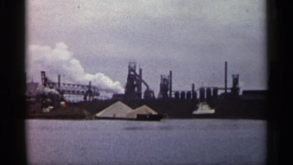 Faded 8mm view of a Michigan industrial complex in 1960, with smoke, vertical stacks, rounded tanks, and water filling the lower frame.