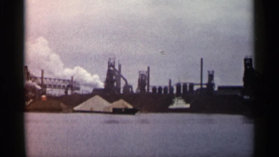 1960 Michigan 8mm frame showing the industrial complex from a slightly shifted angle, with smoke at left and tanks and stacks across the horizon.