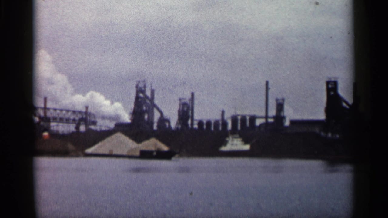 1960 Michigan 8mm frame showing an industrial complex across water, with smokestacks, towers, smoke, pale piles, and a small boat.