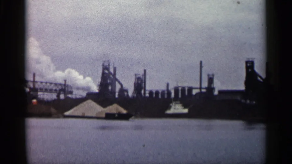 1960 Michigan 8mm frame showing an industrial complex across water, with smokestacks, towers, smoke, pale piles, and a small boat.