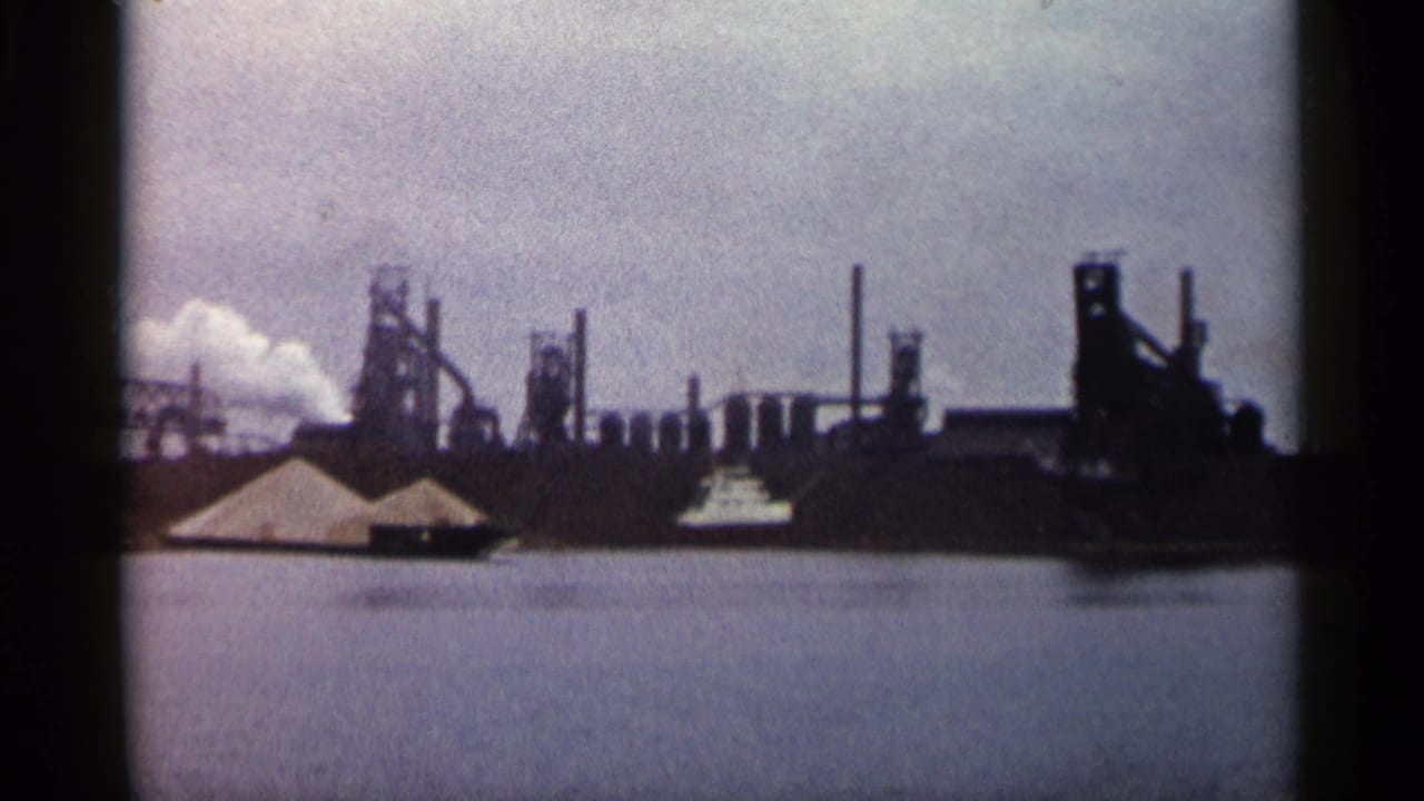Factory skyline across water with smokestacks, smoke, pale piles, and a small boat.