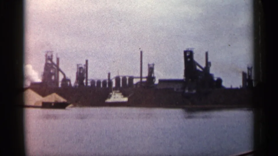 Soft 8mm frame of a Michigan industrial shoreline, showing smokestacks, factory structures, smoke, water, and a small boat near center.