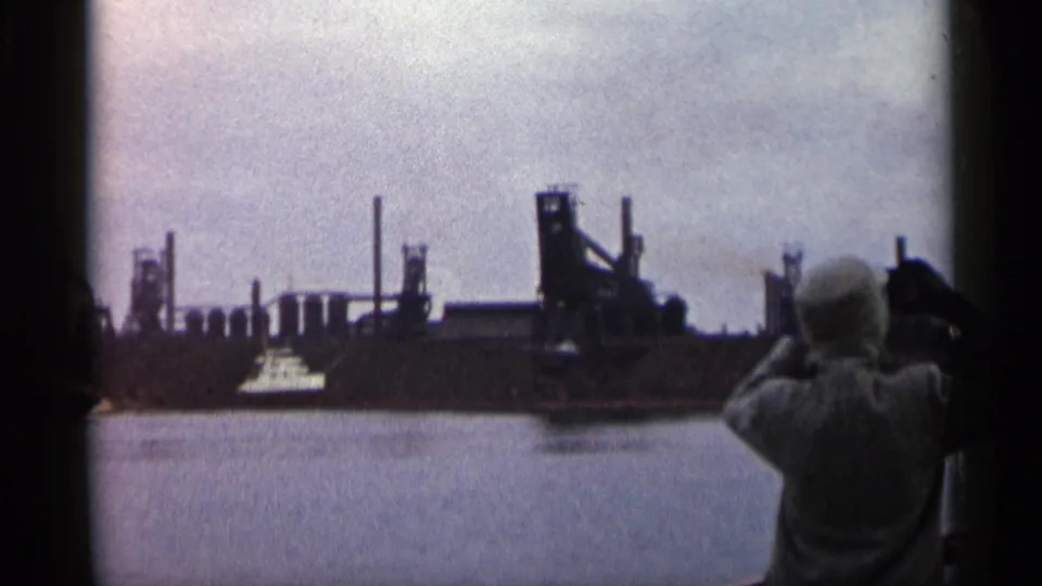 1960 Michigan 8mm frame with a foreground observer facing a waterfront industrial complex, smokestacks, smoke, and a small boat.