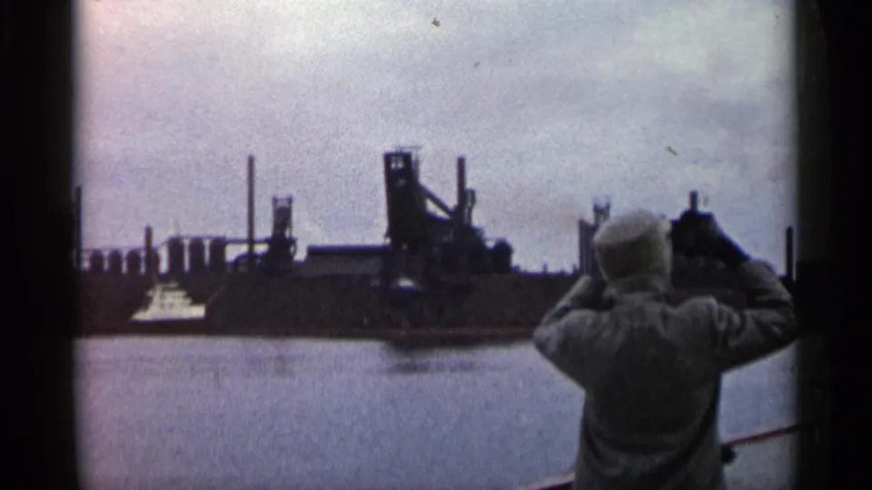 In 1960 Michigan home-movie footage, a woman seen from behind films a dark shipyard scene across harbor water.