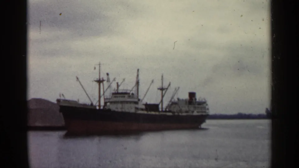 A red and black cargo ship remains in view on Michigan harbor water, captured with faded 8mm color and visible film grain.