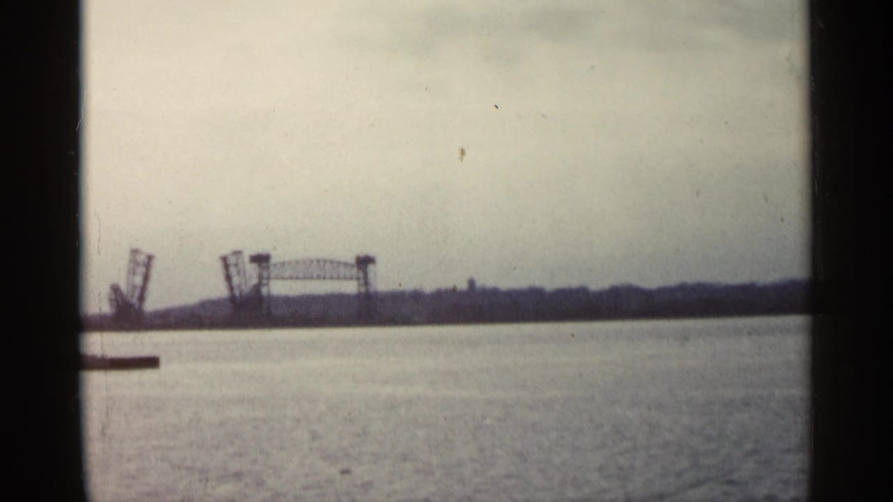 1960 Michigan 8mm frame showing a raised drawbridge in the distance across calm water beneath an overcast sky.