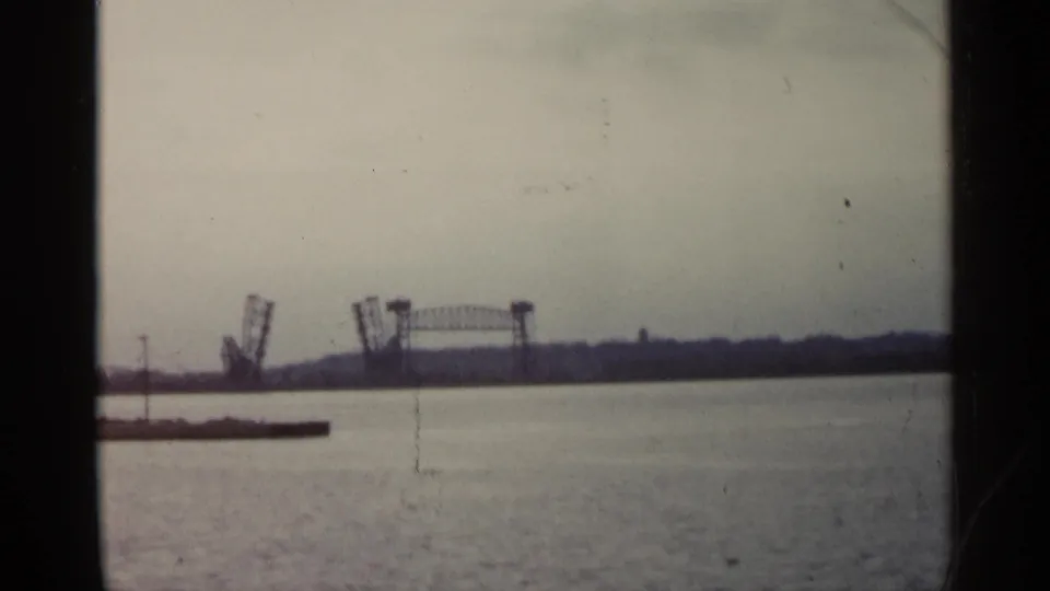 Distant raised drawbridge on a Michigan waterfront, filmed over calm water on faded 8mm stock in 1960.