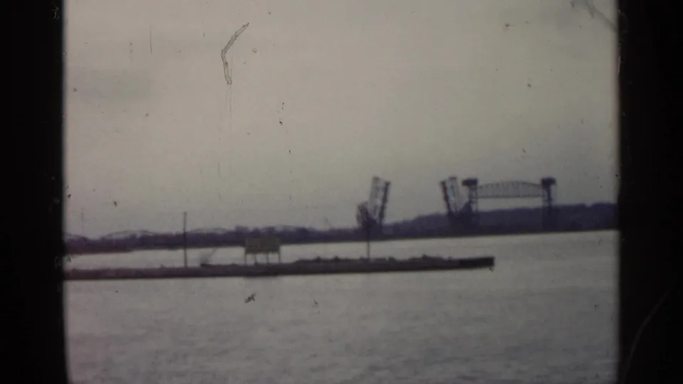 Distant Michigan drawbridge remains raised behind a low waterfront structure, with calm water and cloudy sky visible.