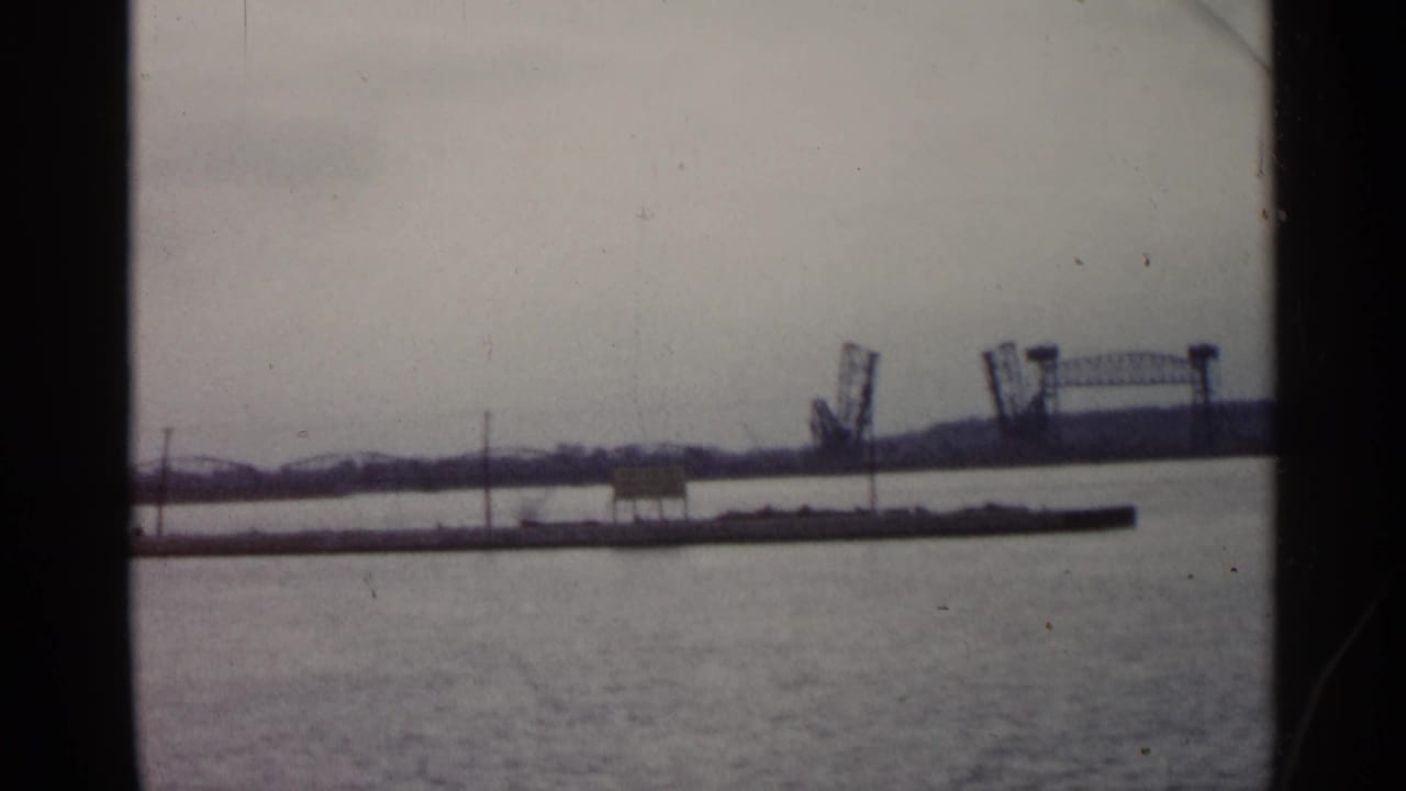1960 Michigan 8mm frame looking across calm water toward a distant bridge-like structure and low shoreline under a pale sky.