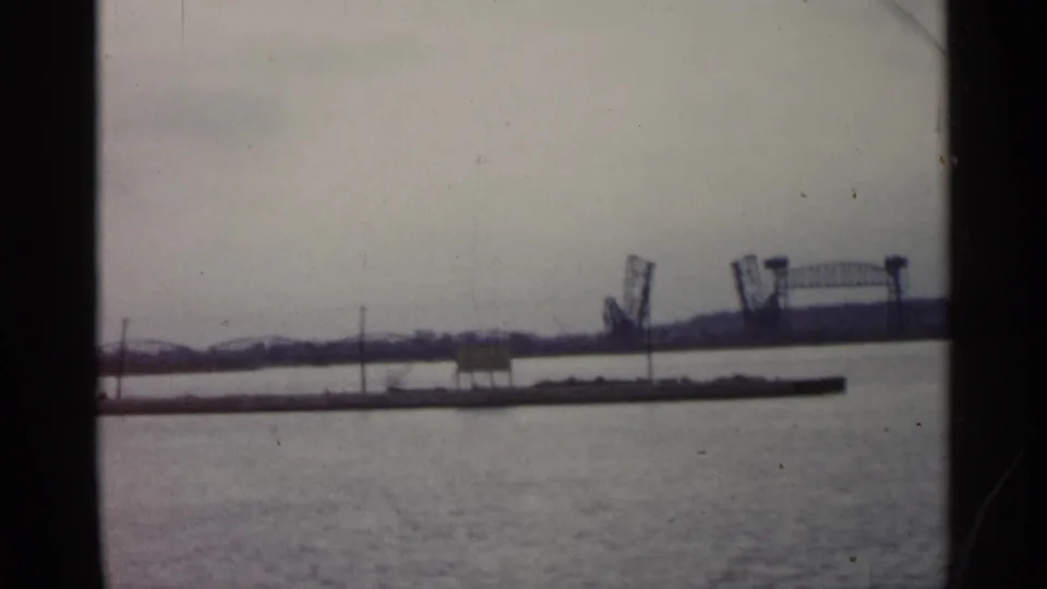 1960 Michigan 8mm frame looking across calm water toward a distant bridge-like structure and low shoreline under a pale sky.