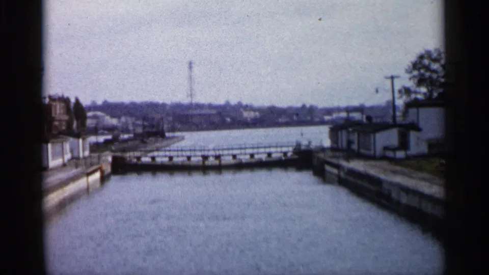 1960 Michigan 8mm view down a quiet canal or lock approach with shoreline walls, a low crossing, distant buildings, and utility poles.