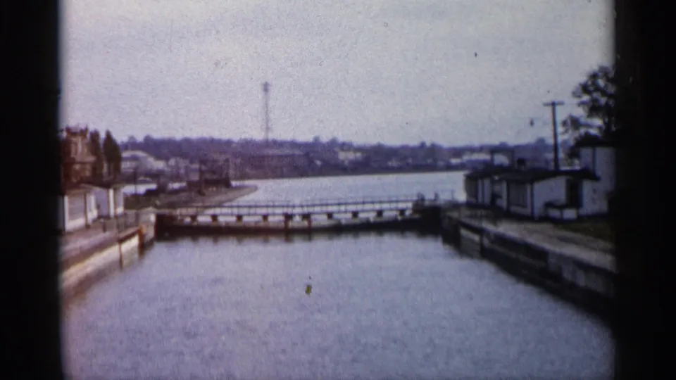 1960 Michigan home-movie frame showing a calm waterway, low crossing, small waterside building, utility poles, and distant shoreline.