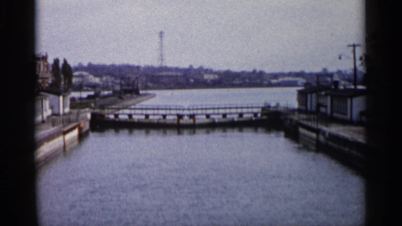 Calm water channel with a low bridge, concrete banks, utility poles, and distant shoreline.