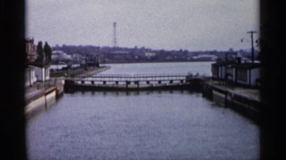 1960 Michigan 8mm frame showing a quiet waterway, low bridge or railing, concrete edges, utility poles, and distant buildings.