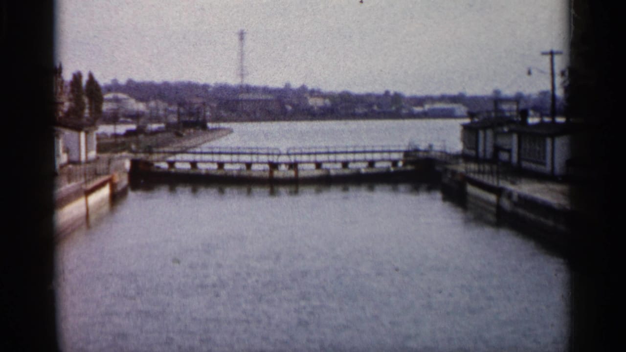 1960 Michigan 8mm view of a water channel, low bridge, concrete waterfront edges, and distant low buildings across the water.