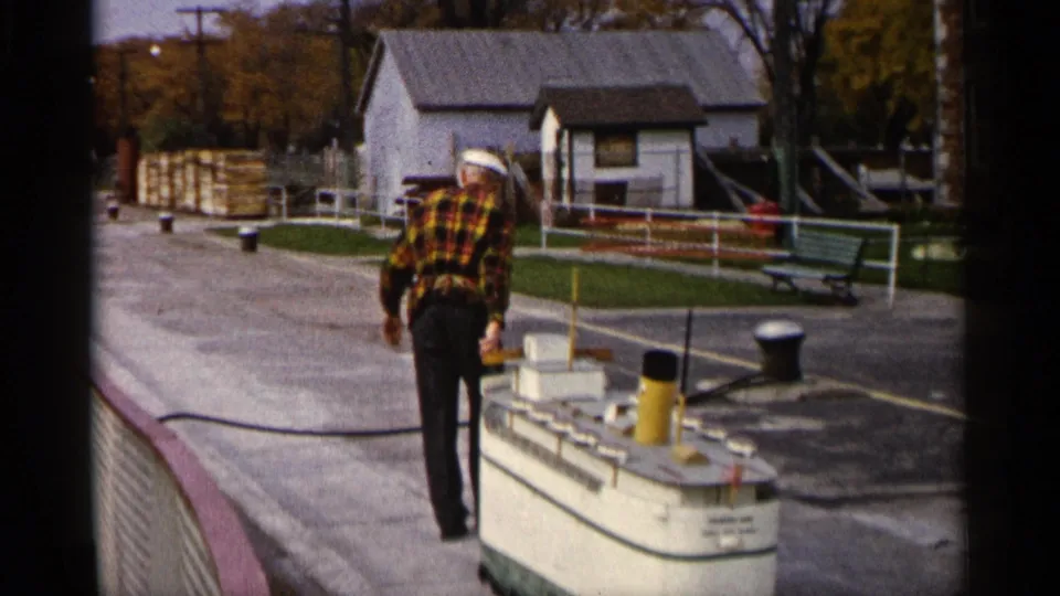 1960 Michigan 8mm frame with a man in plaid walking through a paved dock area near a white building, benches, railings, and bollards.