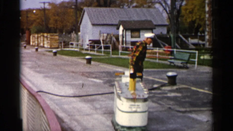 1960 Michigan 8mm frame showing a man near white cylindrical dock equipment, with a small building, benches, railings, and trees behind him.