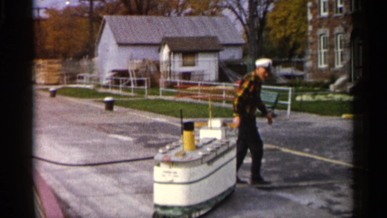 Uniformed man in a white cap stands beside a small model boat on a paved lot.