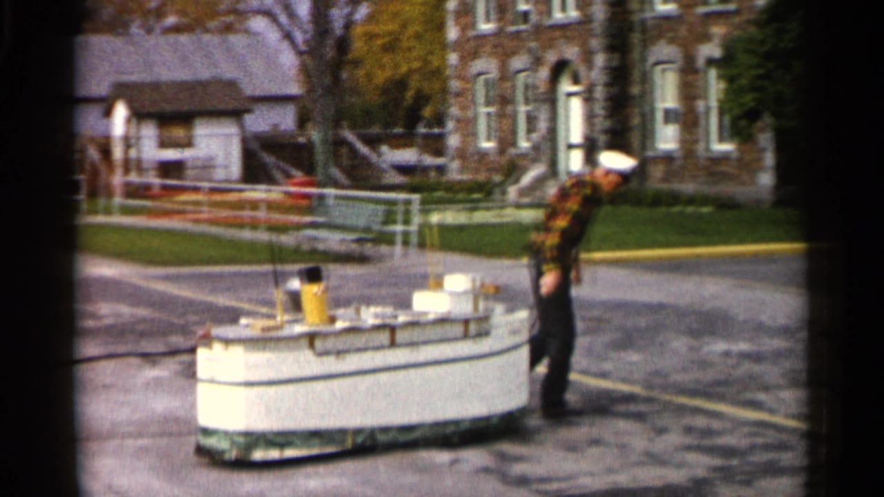 The uniformed man leans over the model vessel on a paved Michigan lot, with a stone building, small shed, railing, and trees behind him.