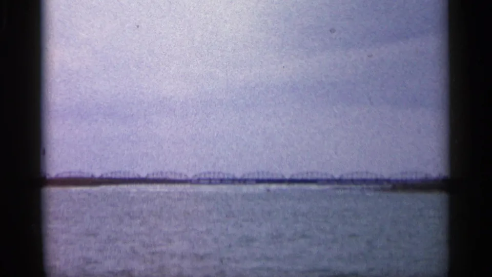 A distant long bridge crosses a body of water in the same 1960 Michigan 8mm clip, seen through soft film grain and dark frame edges.