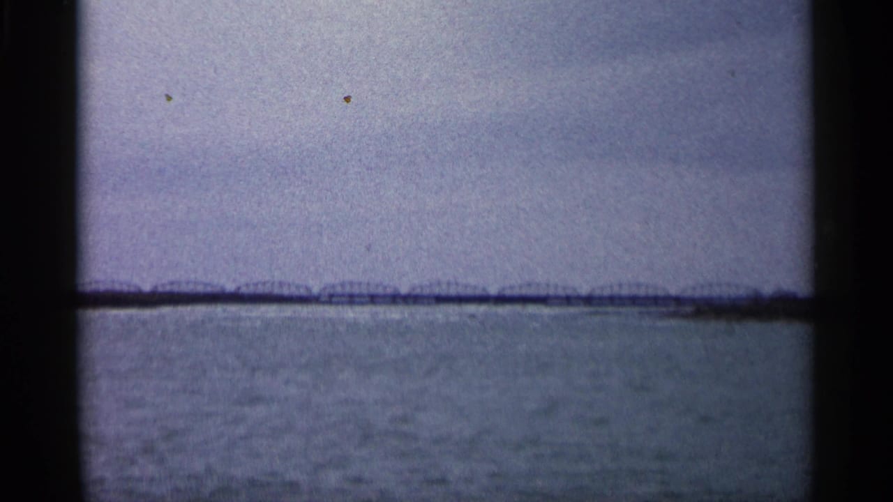 1960 Michigan 8mm frame showing a wide river or lake surface with a distant low bridge or shoreline structure beneath a gray sky.