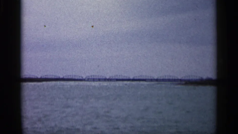 1960 Michigan 8mm frame showing a wide river or lake surface with a distant low bridge or shoreline structure beneath a gray sky.