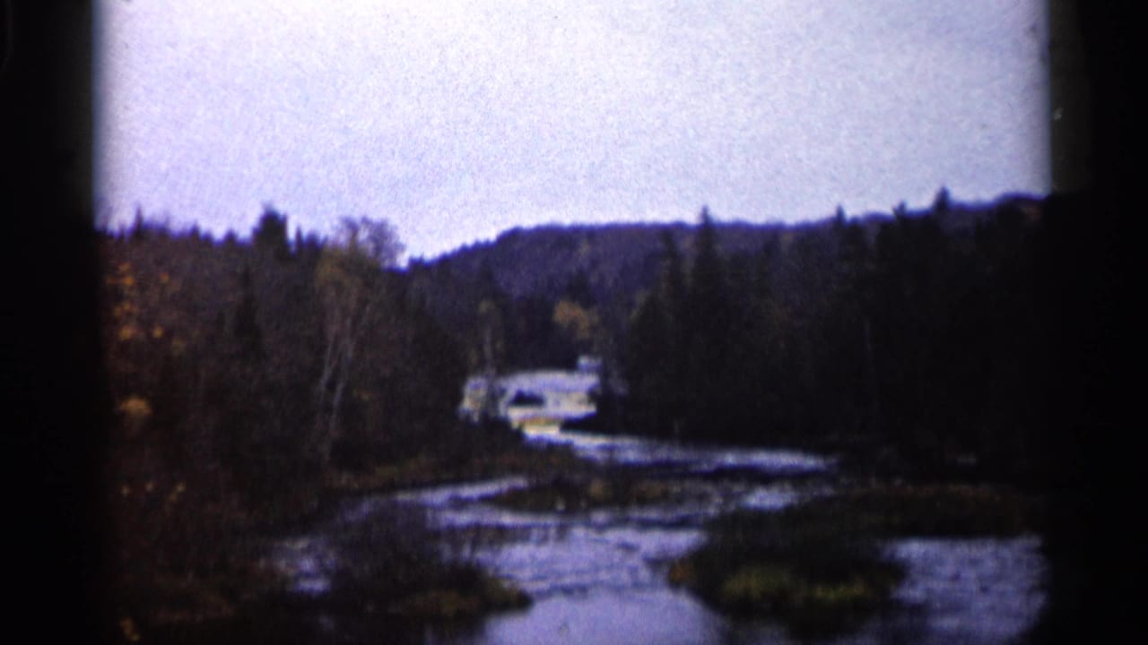 1960 Michigan 8mm frame showing a winding river, coniferous forest, low hills, and a distant light-colored car near the water.