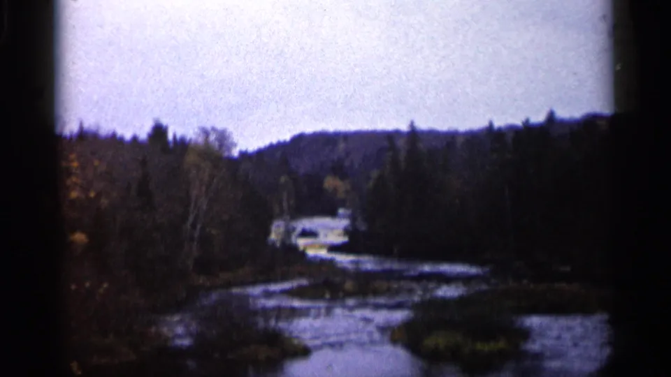 1960 Michigan 8mm frame showing a winding river, coniferous forest, low hills, and a distant light-colored car near the water.