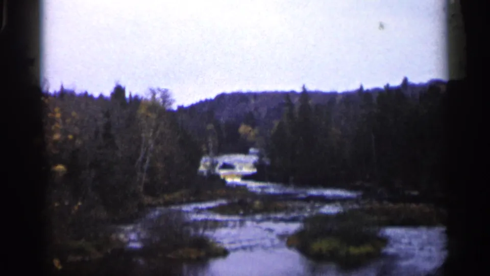 1960 Michigan 8mm frame showing the river channel, mixed forest, low hills, and a distant light-colored car near the far bank.