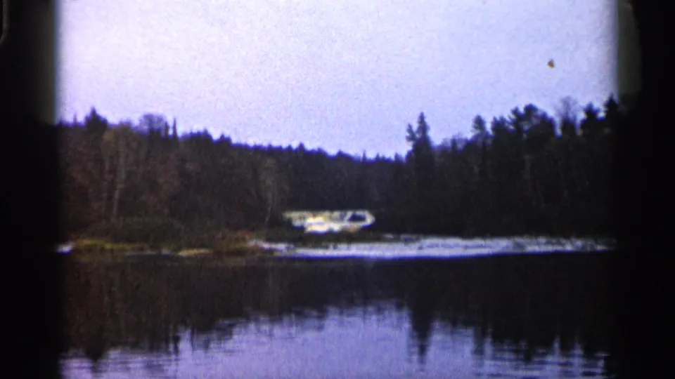 1960 Michigan frame showing still water, dark conifer reflections, a wooded shoreline, and a distant light-colored car near the far bank.