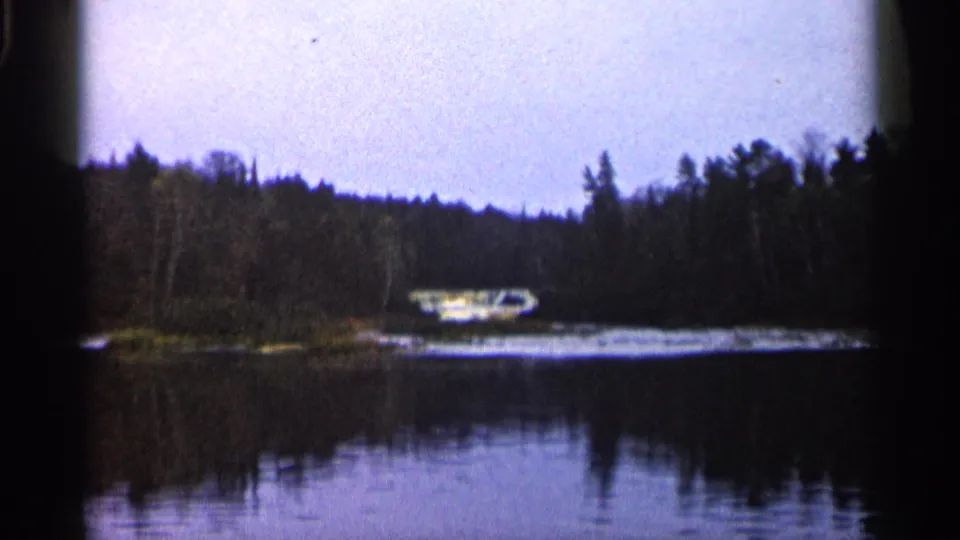 Home-movie frame showing a small lakeside boat below a dark tree line, with calm water in the foreground.