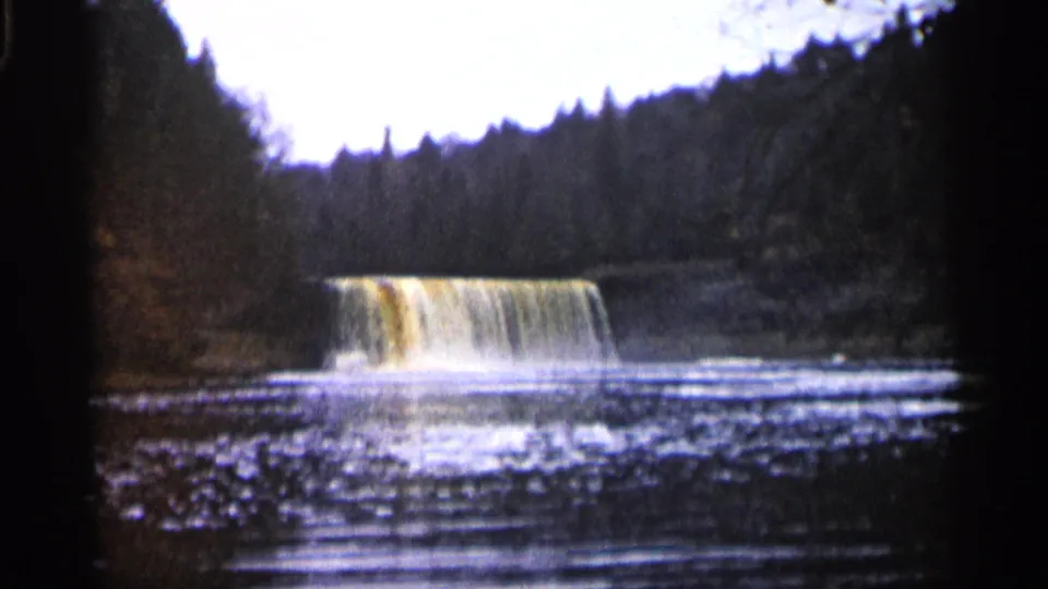 1960 Michigan 8mm frame of a broad waterfall dropping into rippled water, surrounded by wooded banks and evergreens.