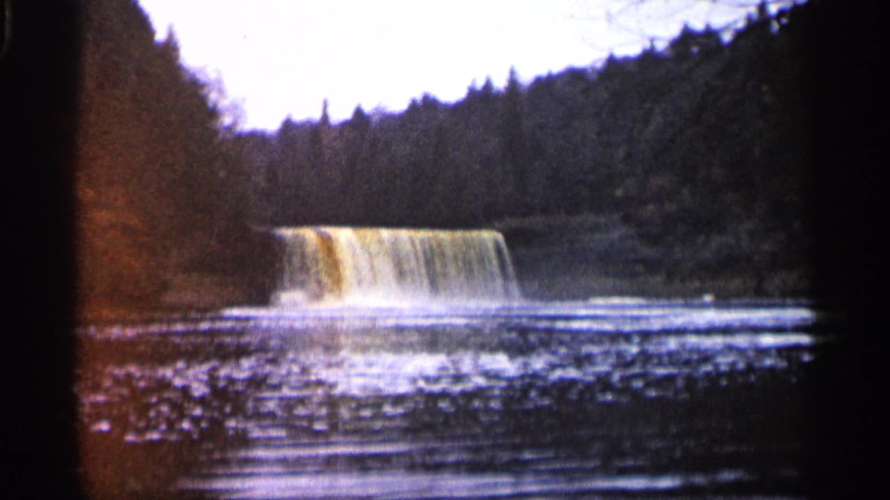 Broad muddy waterfall flowing into a rippling pool below a wooded forest bank.
