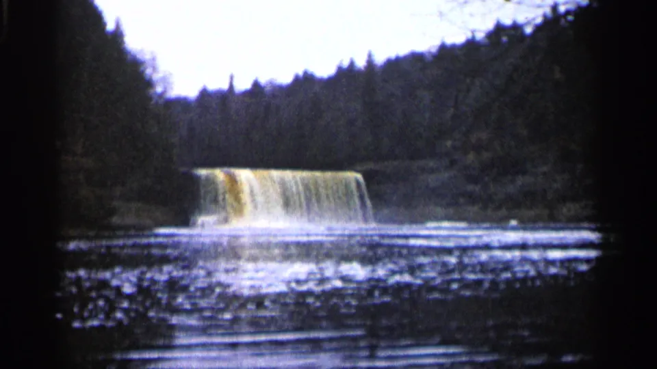 1960 Michigan 8mm view of sediment-colored water dropping over a low ledge into a rippling forest pool.
