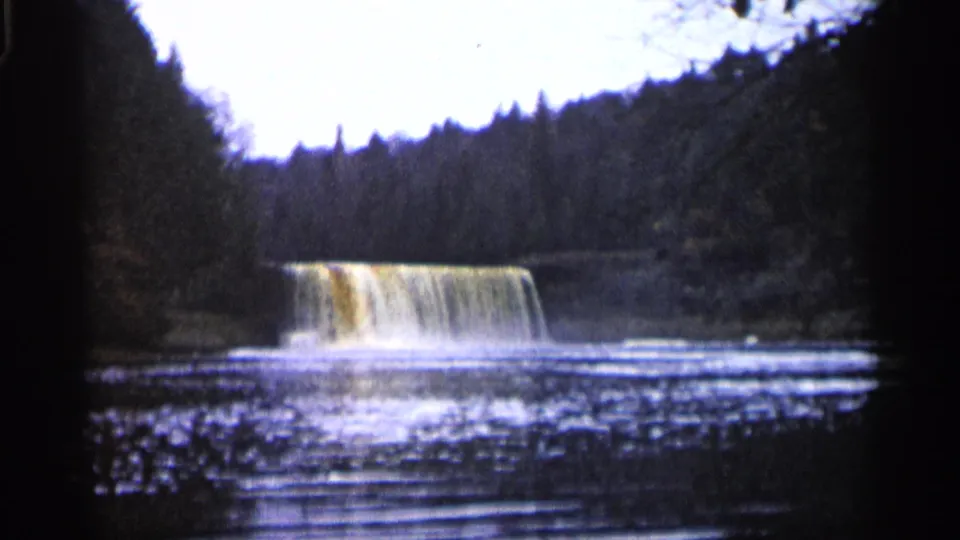 Vintage 8mm frame from Michigan showing a broad forest waterfall and rippling pool with visible grain and dark edge shadows.