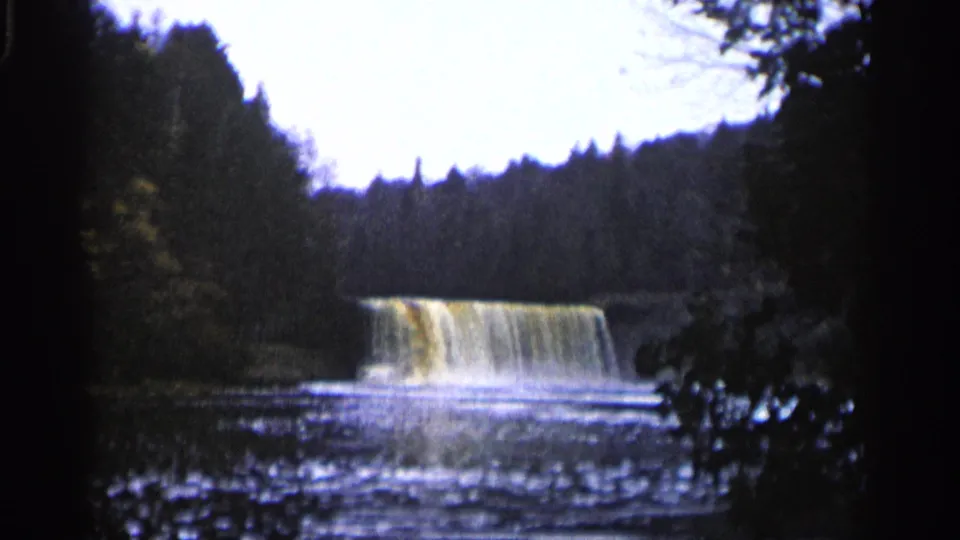 1960 Michigan home-movie frame of a muddy waterfall partly framed by dark trees, branches, and a rippling pool.