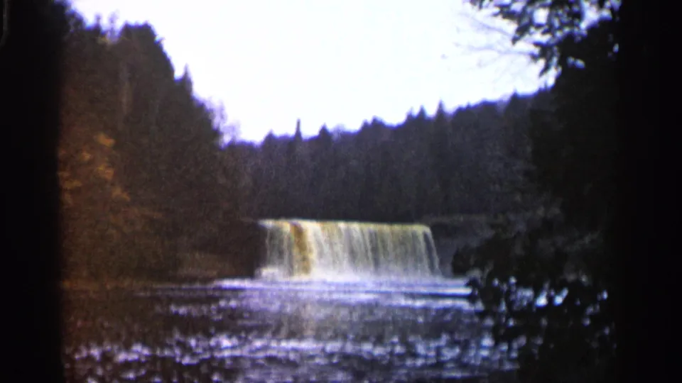 1960 Michigan 8mm frame of a low waterfall and rippled stream seen through dark trees in a forest setting.