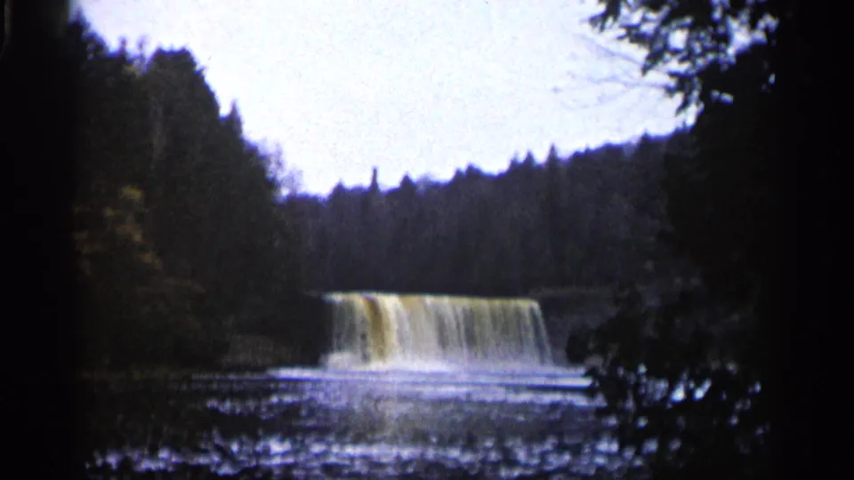 1960 Michigan 8mm view of a waterfall over a low dam or ledge, with forested banks and dark tree silhouettes.