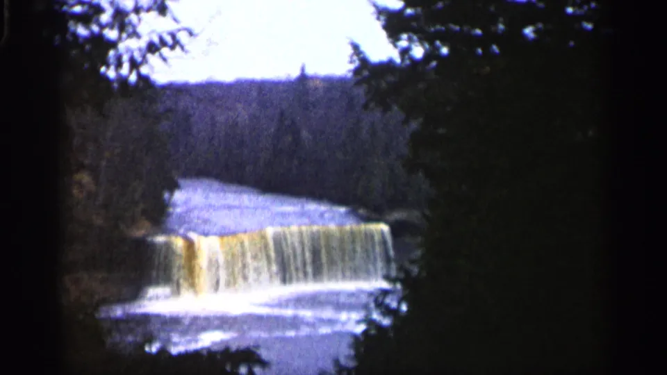 1960 Michigan 8mm frame showing a low waterfall, bright foam, dark trees, and distant wooded terrain.