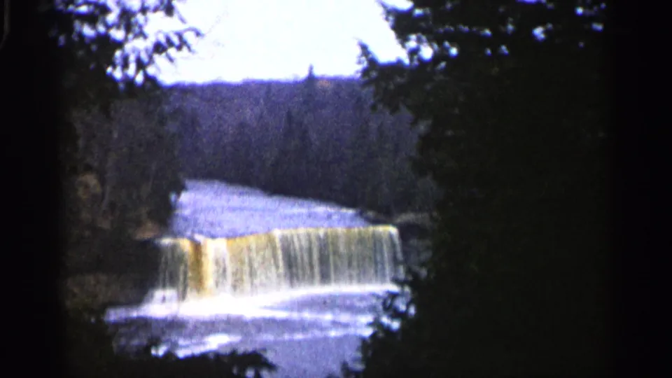 1960 Michigan 8mm frame of a waterfall viewed from a shaded overlook, with river water and wooded hills beyond.
