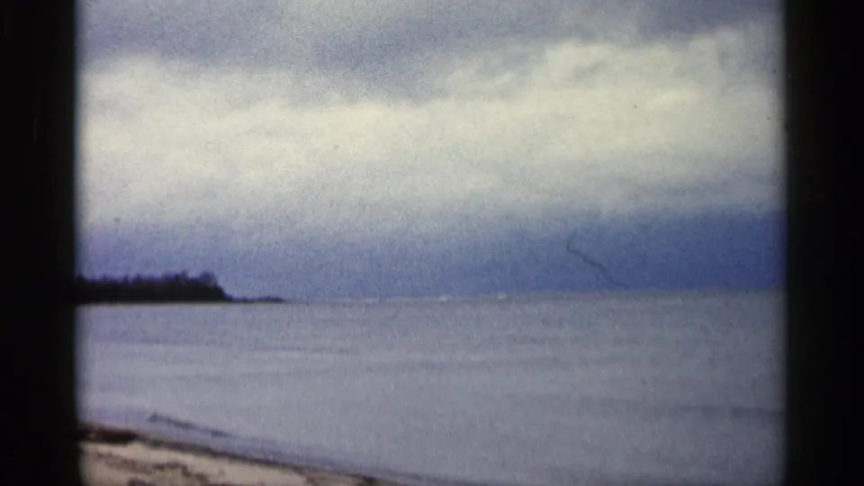 1960 Michigan 8mm frame showing a broad lake horizon, overcast sky, distant point of land, and sandy shore.