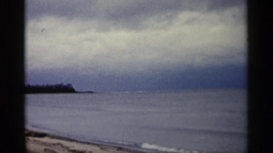 1960 Michigan 8mm frame of a calm lake shore with an overcast horizon and a dark wooded point in the distance.