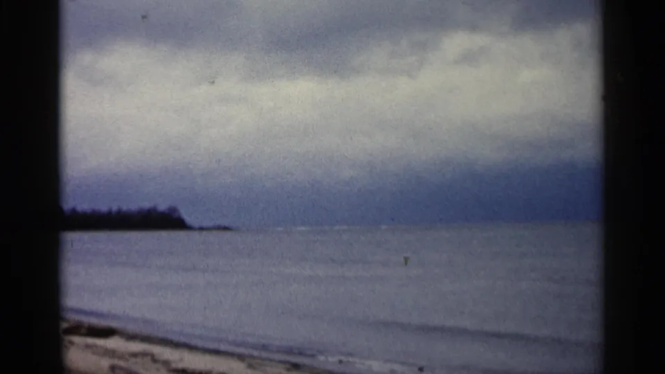1960 Michigan 8mm frame showing calm lake water, sandy beach edge, distant treeline, and heavy overcast sky.