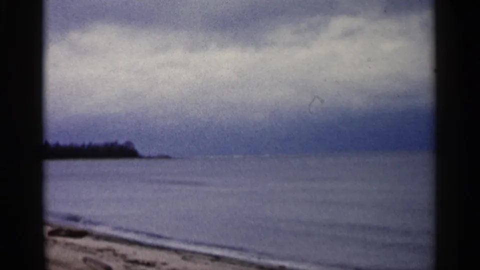 1960 Michigan 8mm frame of a quiet beach edge, smooth lake surface, distant trees, and layered gray-blue clouds.
