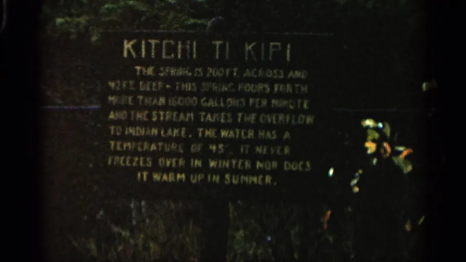 1960 Michigan 8mm frame showing a dark Kitchi Ti Kipi informational sign with yellow text and foliage above.