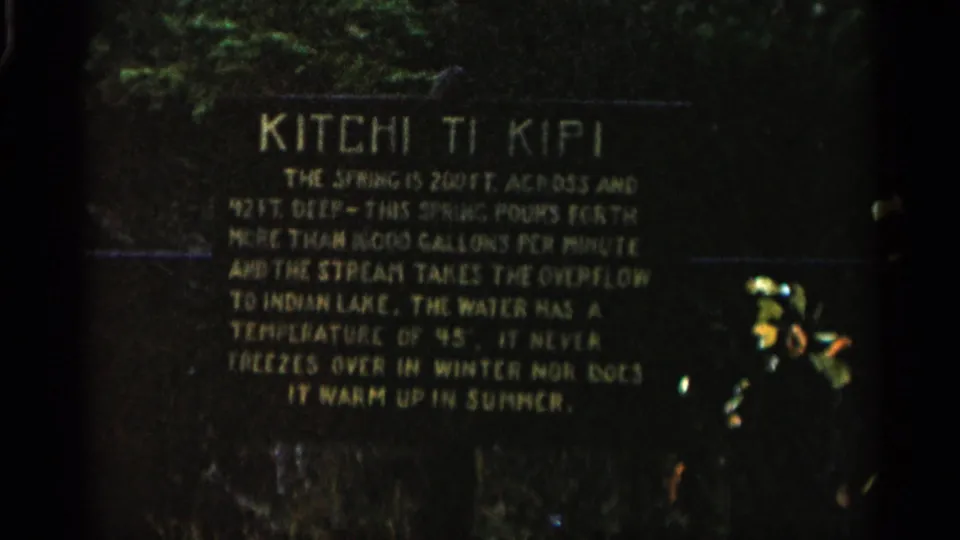 1960 Michigan home-movie frame of the Kitchi Ti Kipi sign, with yellow text describing the spring on a dark background.
