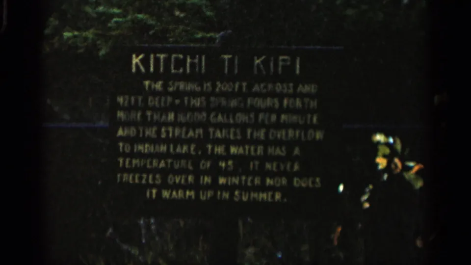 1960 Michigan 8mm frame showing the Kitchi Ti Kipi sign with softened lettering, surrounding foliage, and heavy film grain.