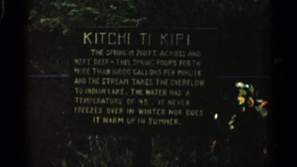 1960 Michigan home-movie frame of the Kitchi Ti Kipi informational board, with grain, low exposure, and sign text still legible.