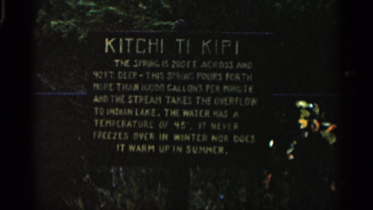 1960 Michigan 8mm frame showing a dark Kitchi Ti Kipi informational plaque with pale lettering and greenery behind it.